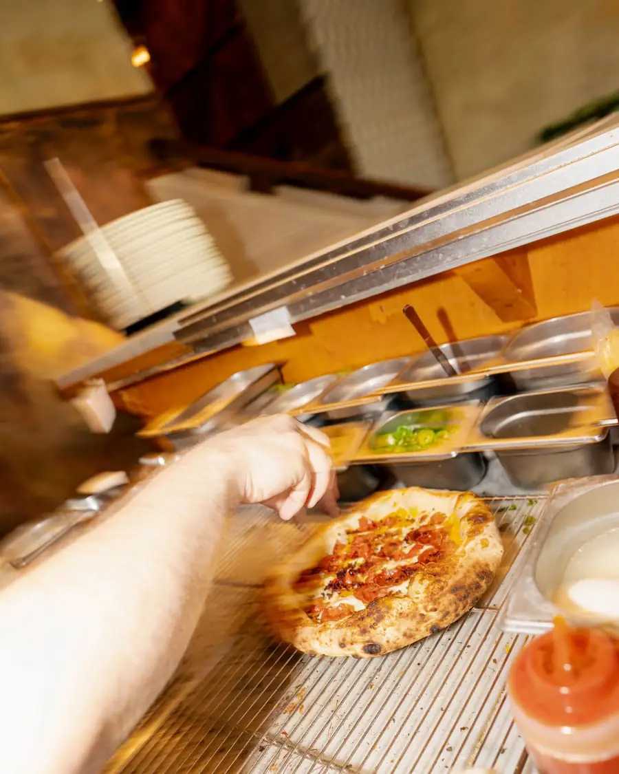 Ręka odstawia pizzę na wydawkę, kuchnia w ruchu